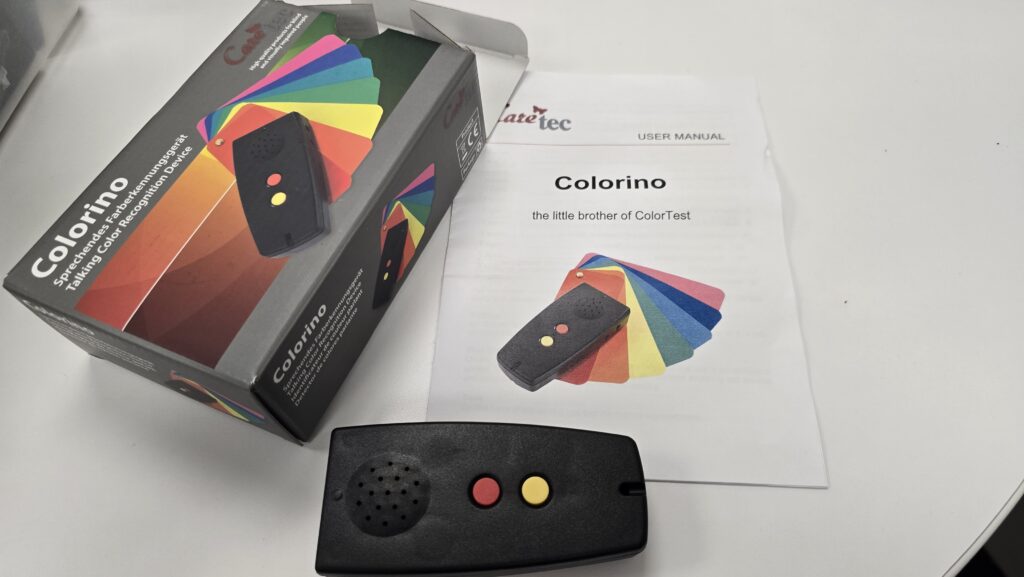 A Colorino talking color recognition device is shown on a white surface, along with its packaging and user manual. The device is black with a speaker grill on the left and two round buttons—one red and one yellow—on the right. The box features the same device image over a fan of colorful swatches, with the text in German and English reading “Colorino Sprechendes Farberkennungsgerät / Talking Color Recognition Device.” The user manual has a similar image and reads “Colorino: the little brother of ColorTest” with the manufacturer name “CareTec” at the top.