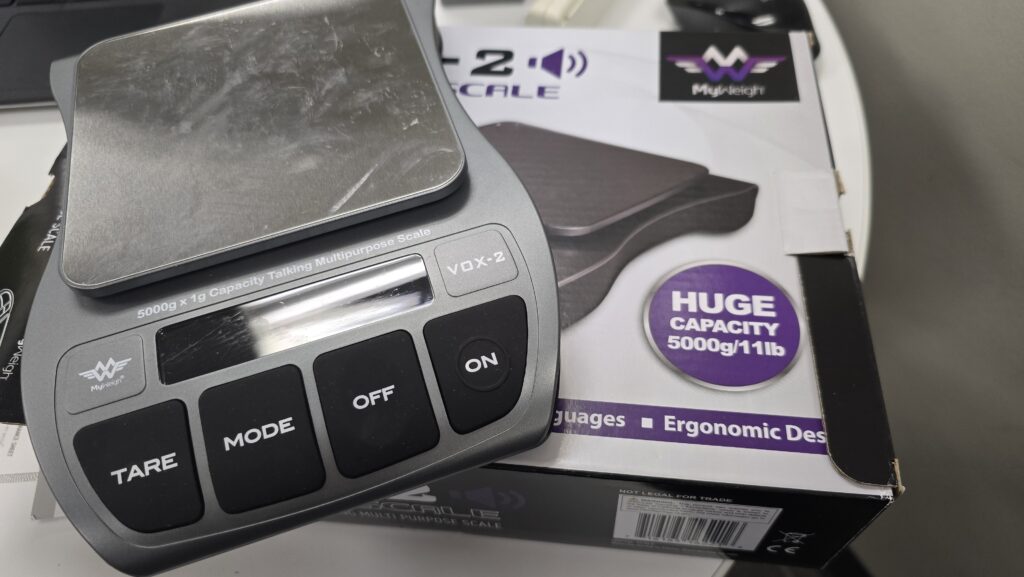 My Weigh VOX-2 talking digital scale with four large buttons labeled Tare, Mode, Off, and On. It has a stainless steel platform and a display screen. The box beneath it shows the scale has a 5000 gram or 11 pound capacity.