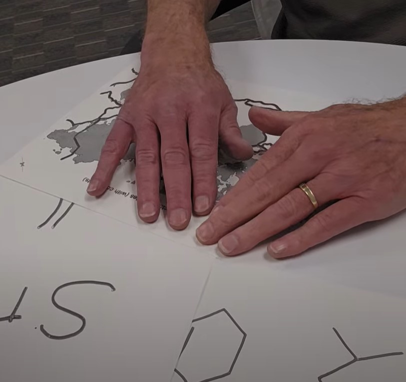 Close-up of a person's hands exploring raised-line tactile graphics on paper, including a tactile map and simple line diagrams. The person is wearing a gold wedding band and is seated at a round white table with several swell-form graphics spread out